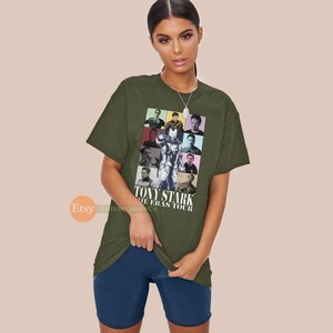 May include: A green t-shirt featuring a collage of images of Tony Stark from the Iron Man films. The text "Tony Stark The Eras Tour" is printed on the shirt.