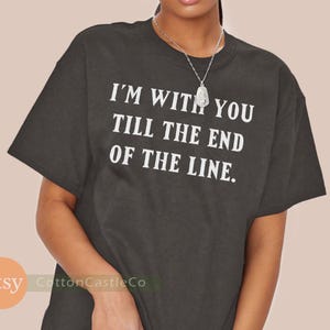May include: Dark gray t-shirt with the white text "I'M WITH YOU TILL THE END OF THE LINE." The shirt is a crew neck style. A silver necklace is visible.