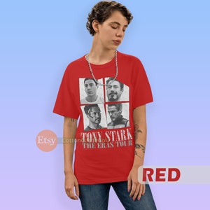 以下が含まれることがあります： アイアンマン映画のトニー・スタークの異なる4つの画像のグラフィックがプリントされた赤いTシャツ。Tシャツには「トニー・スターク・ザ・エラス・ツアー」の文字がプリントされています。