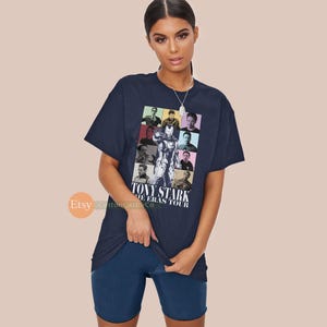 May include: A navy blue t-shirt featuring a colourful graphic of Tony Stark from the Iron Man films. The graphic is a collage of different images of Tony Stark in various poses and outfits. The text "Tony Stark The Eras Tour" is printed below the graphic.
