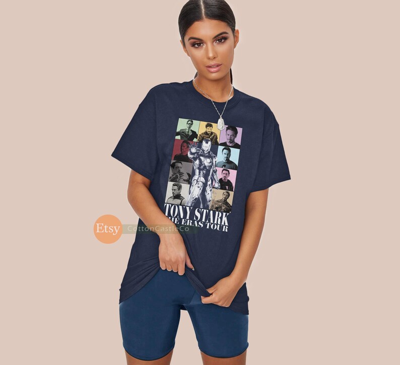 May include: A navy blue t-shirt featuring a colourful graphic of Tony Stark from the Iron Man films. The graphic is a collage of different images of Tony Stark in various poses and outfits. The text "Tony Stark The Eras Tour" is printed below the graphic.
