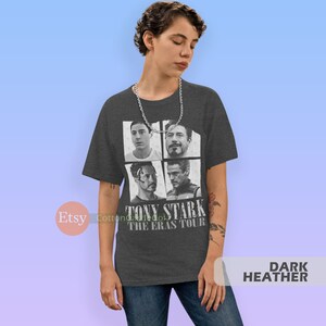 以下が含まれることがあります： ダークヘザーグレーのTシャツに、アイアンマン映画のトニー・スタークの画像4枚のコラージュがプリントされています。Tシャツには「トニー・スターク・ザ・エラス・ツアー」の文字が白でプリントされています。