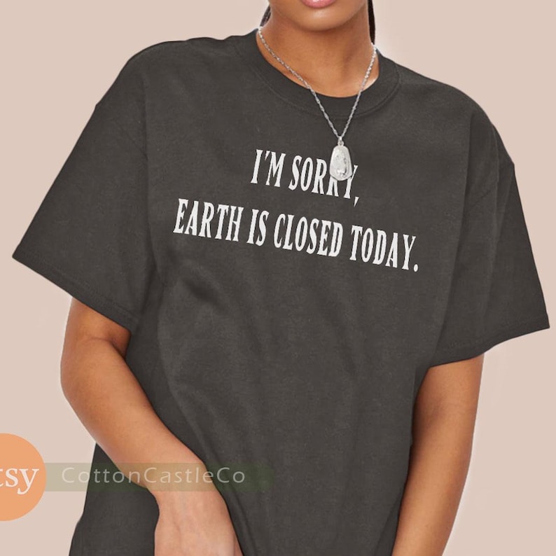 May include: Dark gray t-shirt with the text "I'M SORRY, EARTH IS CLOSED TODAY" in white. The shirt has short sleeves and a crew neck. A silver necklace is visible.