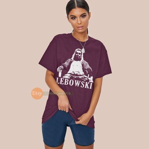 Peut inclure: T-shirt bordeaux avec un graphique blanc d'un homme assis sur un tr&ocirc;ne avec une boisson &agrave; la main. Le texte "The Dude Abides" est imprim&eacute; sous le graphique.