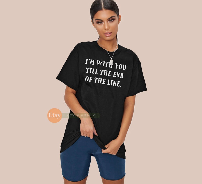 May include: Black t-shirt with the text "I'M WITH YOU TILL THE END OF THE LINE" in white. The shirt is paired with dark blue shorts. The model is wearing a silver necklace.