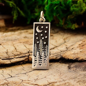 Puede incluir: Un dije de plata con un diseño detallado de una luna creciente, estrellas y pinos sobre un fondo oscuro. El dije es rectangular y tiene un bucle en la parte superior para sujetarlo a una cadena.