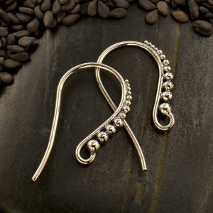 May include: A pair of silver ear climbers. Each earring has a curved design with small, round beads along the curve. The earrings are displayed on a dark, textured surface, contrasting with the bright metal. The earrings are a type of jewellery.