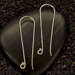 Puede incluir: Un par de pendientes plateados de diseño minimalista. Cada pendiente tiene un alambre largo y recto que se curva en la parte superior y termina en una pequeña bola redondeada. Los pendientes se exhiben sobre una piedra negra lisa, rodeados de semillas negras.