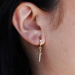 Puede incluir: Aretes de aro dorados con un dije de daga de plata que cuelga de la parte inferior.