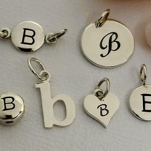 May include: A collection of silver-toned letter "B" charms in various shapes. Includes a round charm with the letter "B", a heart-shaped charm with "B", and a lowercase "b" charm. The charms are designed for jewelry making.
