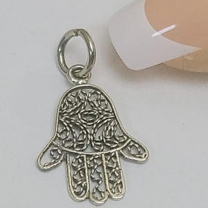 Puede incluir: Un dije de mano de Hamsa plateado con intrincados detalles de filigrana. El dije presenta un diseño de mano abierta con un lazo en la parte superior para sujetar. El fondo es de color claro.