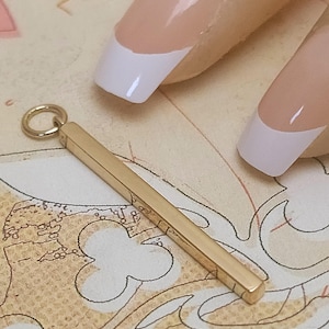 May include: A gold-colored, rectangular pendant with a small loop at one end. The pendant is shown against a patterned background, with a person's nails in the upper right corner.