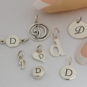 May include: A collection of silver-colored charms, each featuring the letter "D". The charms vary in shape, including round, heart, and a stylized letter "D". Some charms have a loop for attaching to a bracelet or necklace.