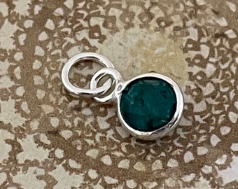 Puede incluir: Un pequeño colgante redondo con una piedra verde oscuro engastada en un metal plateado. El colgante tiene un lazo en la parte superior para sujetarlo a una cadena o pulsera. La piedra es de un tono verde intenso y rico.