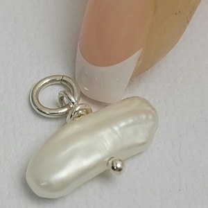 Puede incluir: Un colgante de perla barroca blanca con una anilla de plata y un anillo de plata. La perla está unida al anillo con una cuenta de plata.