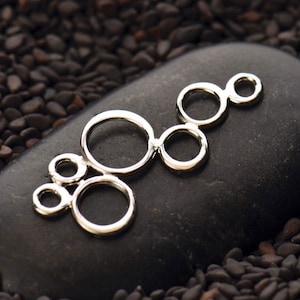 Könnte beinhalten: Silberfarbener Metall-Anhänger mit einem Design aus miteinander verbundenen Kreisen in verschiedenen Größen. Der Anhänger ist ein dekoratives Element für die Schmuckherstellung.