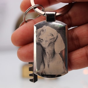 May include: A silver keyring with a rectangular tag featuring a black and white engraved image of a dog's head.