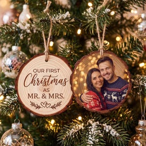Peut inclure: Deux décorations rondes en tranche de bois suspendues à un sapin de Noël. L'une porte l'inscription "OUR FIRST Christmas AS MR. & MRS." avec un motif de cœur. L'autre présente une photographie couleur d'un couple portant des pulls de Noël.