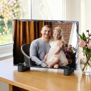 May include: A rectangular photo slate with a rough, chiseled edge featuring a printed image of an adult and a child sitting together. The slate is supported by two black plastic stands, displaying a portrait of a person in a grey jumper holding a child.