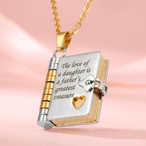 Peut inclure: Un collier doré avec un pendentif en forme de livre. Le pendentif présente des accents argentés et dorés, avec l'inscription "The love of a daughter is a father's greatest treasure." Un petit cœur doré est visible.
