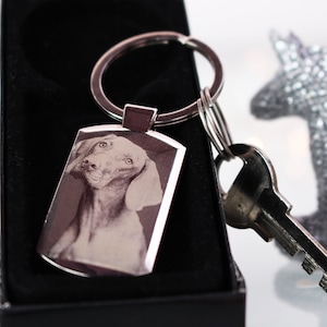 May include: A silver keyring with a black and white photo of a dog engraved on a rectangular tag. The keyring is attached to a silver key ring.