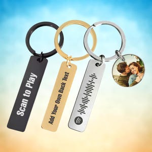 Könnte beinhalten: Drei personalisierte Schlüsselanhänger mit verschiedenen Designs. Ein schwarzer Schlüsselanhänger mit der Aufschrift "Scan to Play", ein goldener Schlüsselanhänger mit der Aufschrift "Add Your Own Back Text" und ein silberner Schlüsselanhänger mit einem Spotify-Code. Ein vierter Schlüsselanhänger hat ein Foto.