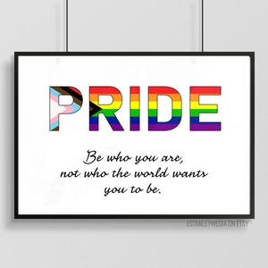 Puede incluir: Una impresión enmarcada en blanco con la palabra "PRIDE" en colores arcoíris. Las letras están formadas por diferentes banderas del orgullo. La cita "Sé quien eres, no quien el mundo quiere que seas." está impresa debajo de la palabra "PRIDE".