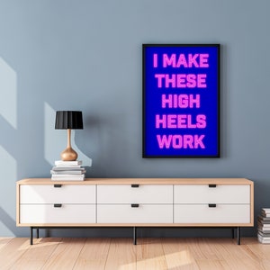 May include: A framed print with a blue background and pink neon text that reads "I MAKE THESE HIGH HEELS WORK". The print is hanging on a light blue wall above a white dresser with six drawers.