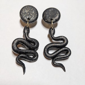 May include: A pair of black snake dangle earrings with gold glitter accents. The earrings are made of polymer clay and have a black glitter finish.