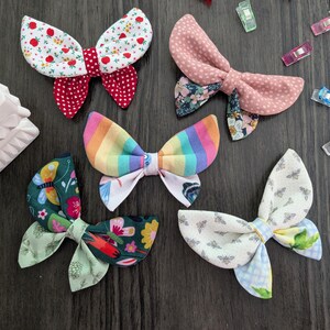 May include: Assortment of handmade fabric hair bows in various patterns and colors, including red polka dots, floral, rainbow stripes, and bees. The bows are arranged on a dark wooden surface.