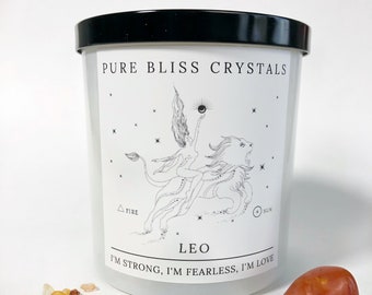 May include: White candle with a black lid and a black and white illustration of a woman riding a lion. The candle has the text "PURE BLISS CRYSTALS" and "LEO I'M STRONG, I'M FEARLESS, I'M LOVE" on the front. There are small orange and white crystals scattered in front of the candle and a large orange crystal to the right.