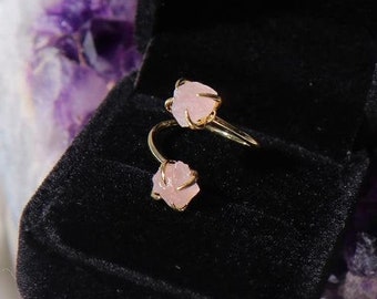 May include: A gold ring with two pink rose quartz stones set in a delicate, curved design. The ring is displayed in a black velvet ring box.
