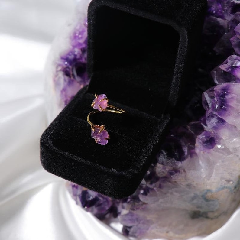 May include: A gold ring with two pink amethyst stones set in the band. The ring is in a black velvet ring box.