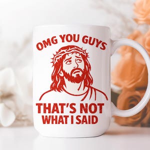Pu&ograve; includere: Tazza in ceramica bianca con grafica rossa di un uomo con corona di spine e la scritta "OMG YOU GUYS THAT'S NOT WHAT I SAID". La tazza ha un manico curvo. Lo sfondo &egrave; sfocato.