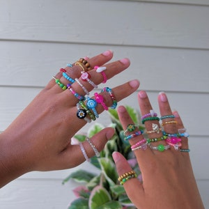 Op de afbeelding: Een handmodel draagt ​​een verscheidenheid aan kleurrijke kralenringen. De ringen zijn gemaakt van verschillende gekleurde kralen, waaronder roze, blauw, groen en wit. Sommige ringen hebben kleine bedels, zoals bloemen en sterren.