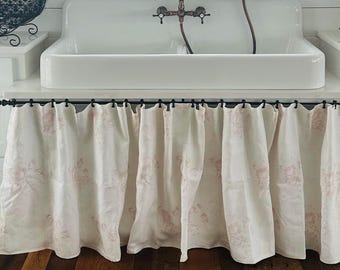 May include: A white farmhouse-style sink with a vintage faucet is the focal point of this kitchen. The sink is set against a backdrop of white cabinets and a window with floral curtains. The floor is made of wood.
