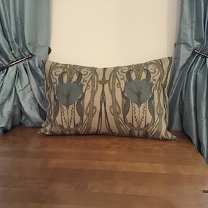 May include: A rectangular cushion featuring a floral pattern in blue, grey, and beige tones. It rests on a wooden surface, flanked by light blue curtains held by gold-coloured metal rings. The design has an Art Nouveau feel.
