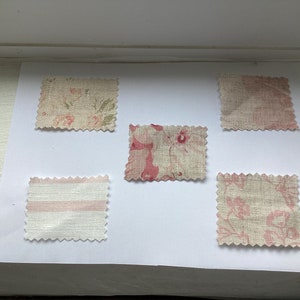 May include: Five fabric samples with pink floral and striped designs. The swatches are displayed on a white surface, highlighting different patterns and textures. The fabrics appear to be linen or a similar material.