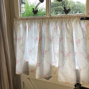 May include: A white curtain with a pink rose pattern, hanging in front of a window. The curtain is made of a light fabric and is gathered at the top. The window reveals a view of green foliage outside.
