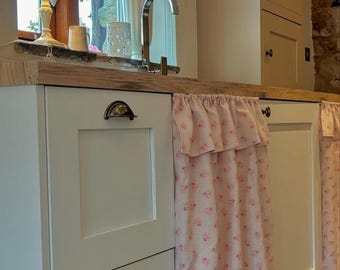 May include: A kitchen interior featuring white cabinets with bronze hardware, a wooden countertop, and a pink floral curtain. A vase of pink roses sits near a window with a decorative valance. A small lamp is on the countertop.