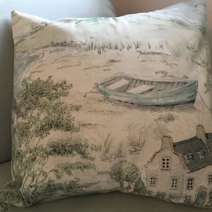 May include: Square throw pillow with a scenic landscape print. The design features a light blue boat on a sandy shore, a small house, and trees. The color palette includes shades of blue, green, and beige.