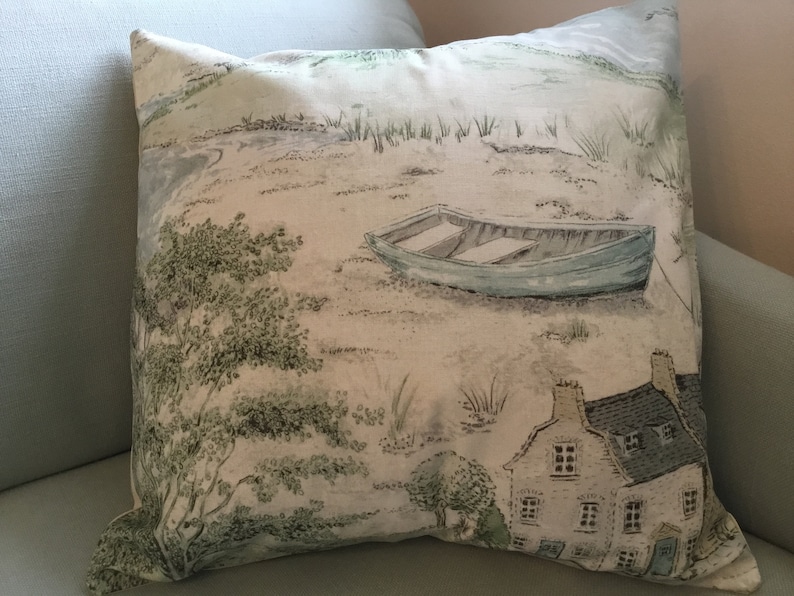 May include: Square throw pillow with a scenic landscape print. The design features a light blue boat on a sandy shore, a small house, and trees. The color palette includes shades of blue, green, and beige.