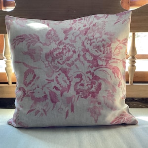 Könnte beinhalten: Quadratisches Zierkissen mit einem rosa Blumenmuster auf hellem Hintergrund. Das Kissen liegt auf einer weißen Oberfläche, im Hintergrund sind eine Holzlehne und ein Fenster zu sehen. Das Design erinnert an Vintage-Stil.