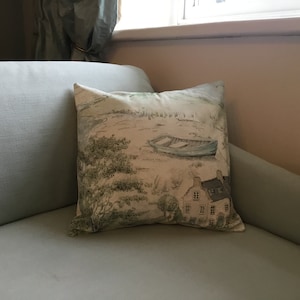 May include: Square throw pillow with a scenic landscape print. The pillow features a light blue boat on a shore, a house, and trees in shades of green and beige. The pillow is resting on a light blue sofa.