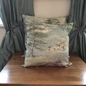 May include: Two square throw pillows with a scenic landscape design in shades of blue, green, and beige. The pillows feature a tree, figures, and a building. The pillows are placed on a wooden surface, with blue curtains in the background.