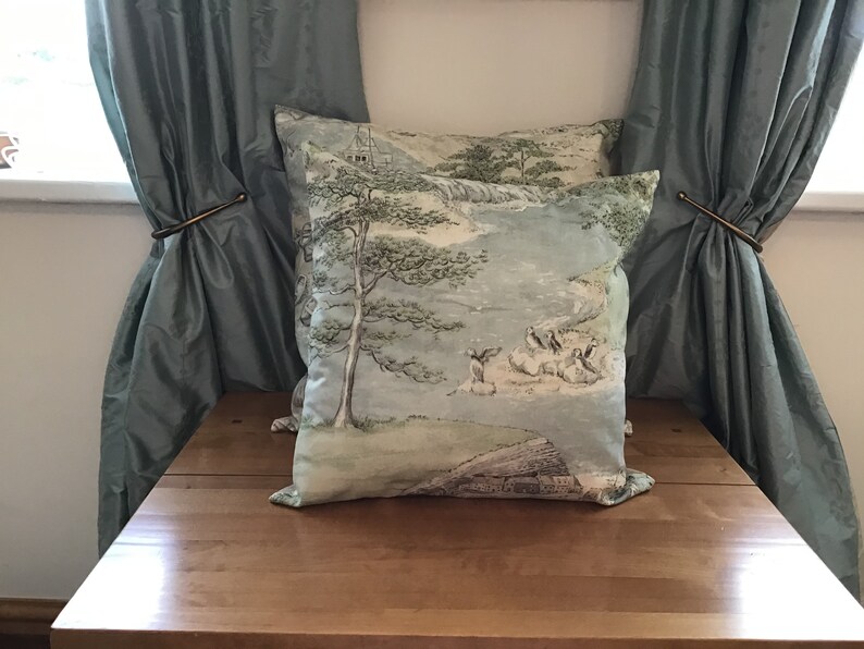 May include: Two square throw pillows with a scenic landscape design in shades of blue, green, and beige. The pillows feature a tree, figures, and a building. The pillows are placed on a wooden surface, with blue curtains in the background.