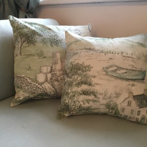 May include: Two square throw pillows with a pastoral landscape design. The pillows feature a light green and white color scheme with illustrations of a castle, trees, a boat, and a small house. The pillows are placed on a light blue sofa.