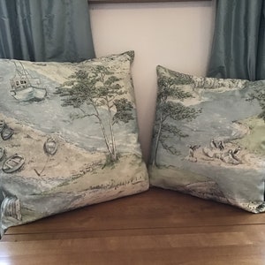 May include: Two square throw pillows with a coastal scene design. The pillows feature a light blue and green color palette with illustrations of boats, trees, and a shoreline. The pillows are placed on a wooden surface with blue curtains in the background.