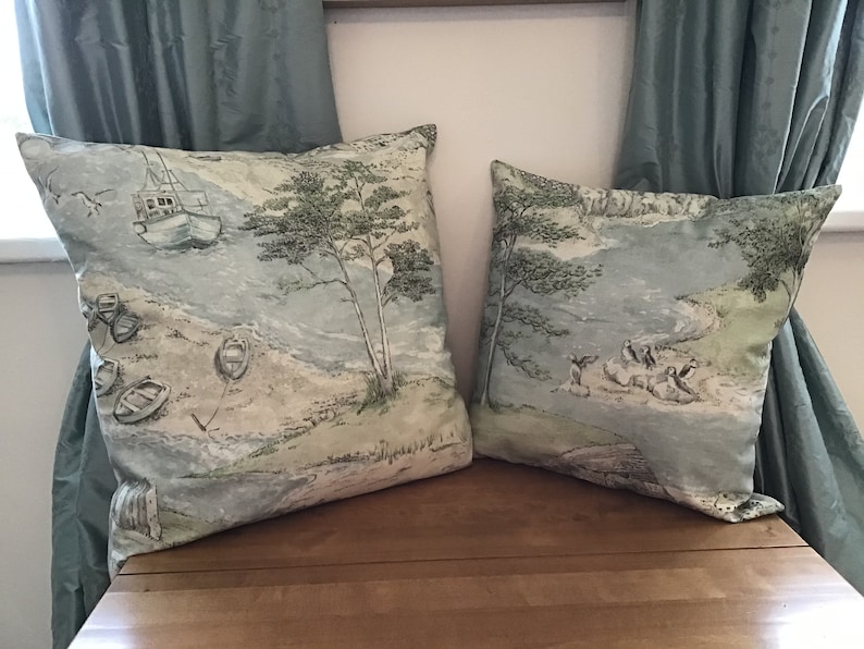 May include: Two square throw pillows with a coastal scene design. The pillows feature a light blue and green color palette with illustrations of boats, trees, and a shoreline. The pillows are placed on a wooden surface with blue curtains in the background.