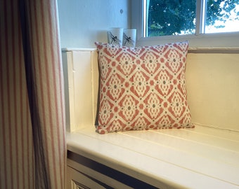 May include: A square decorative pillow with a repeating geometric pattern in red, white, and gray. The pillow is placed on a white bench near a window. A striped curtain is visible on the left.